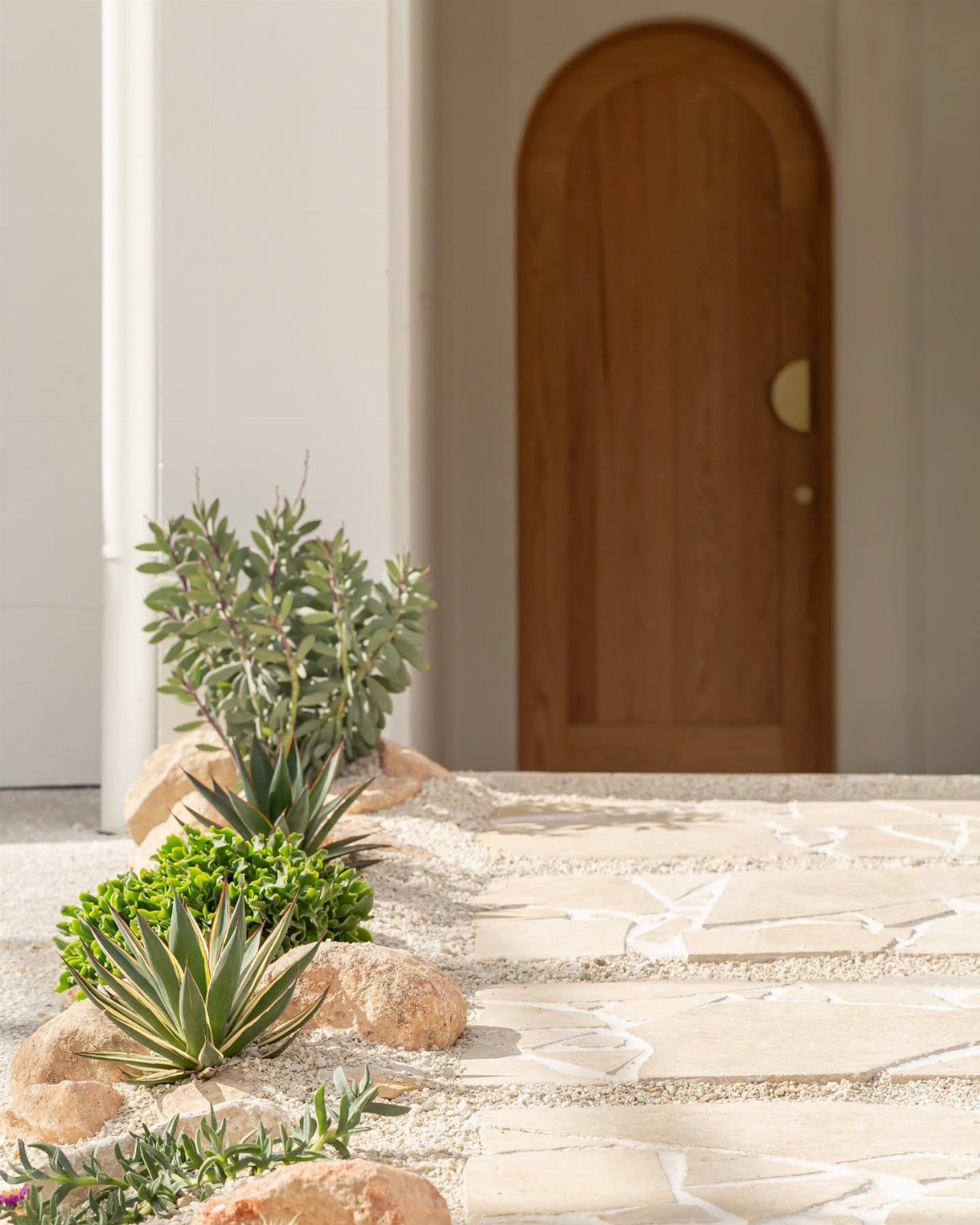 Irregular stone flagstone pathway bordered by desert plants and large rocks leading to an arched wooden door.