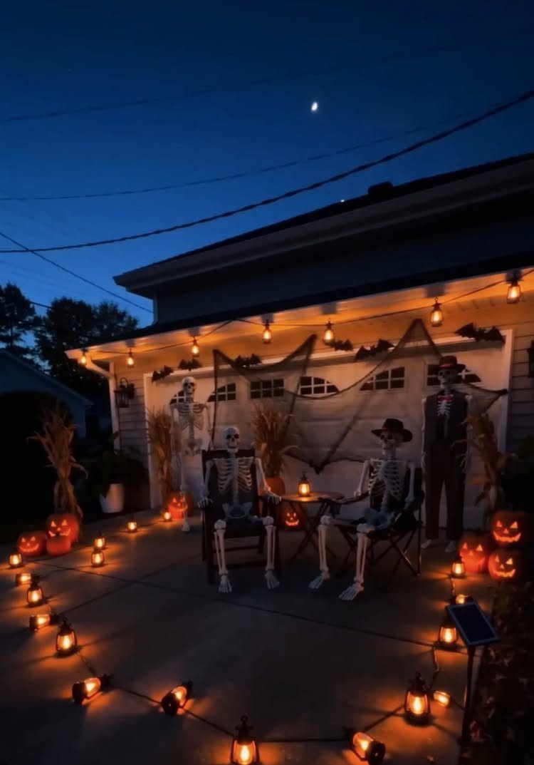 Skeletons sitting in chairs on a driveway decorated with glowing Halloween lanterns and string lights.
