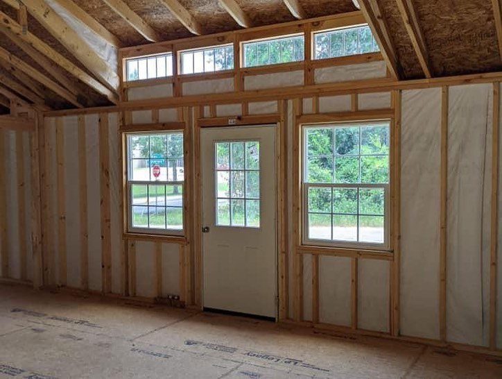 Unfinished interior of a wooden shed house showing studs, insulation, a white door, and windows.
