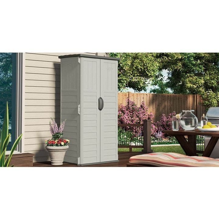 A grey resin vertical storage shed stands on a wooden deck next to a white house.