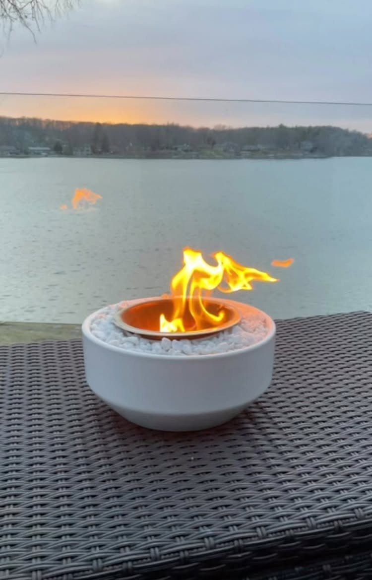 A small white tabletop fire pit burning with bright orange flames on a wicker table by a lake.