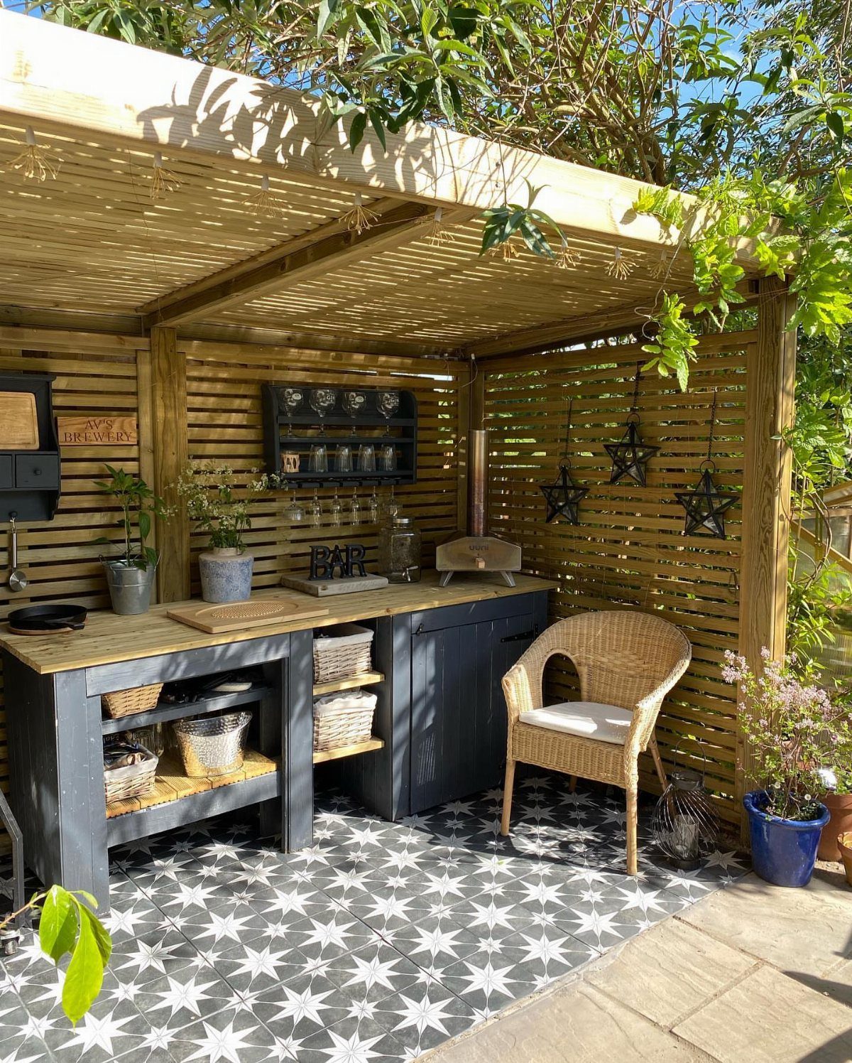 10 Wooden Garden Kitchen Ideas for Warm Outdoor Cooking 4 Outdoor kitchen setup featuring dark grey cabinetry and decorative star-patterned floor tiles under a wooden pergola.