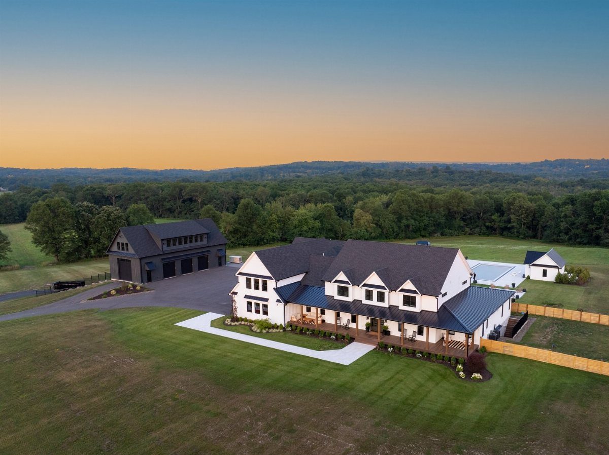 A white traditional farmhouse with a dark roof and a large detached garage on a green lawn.