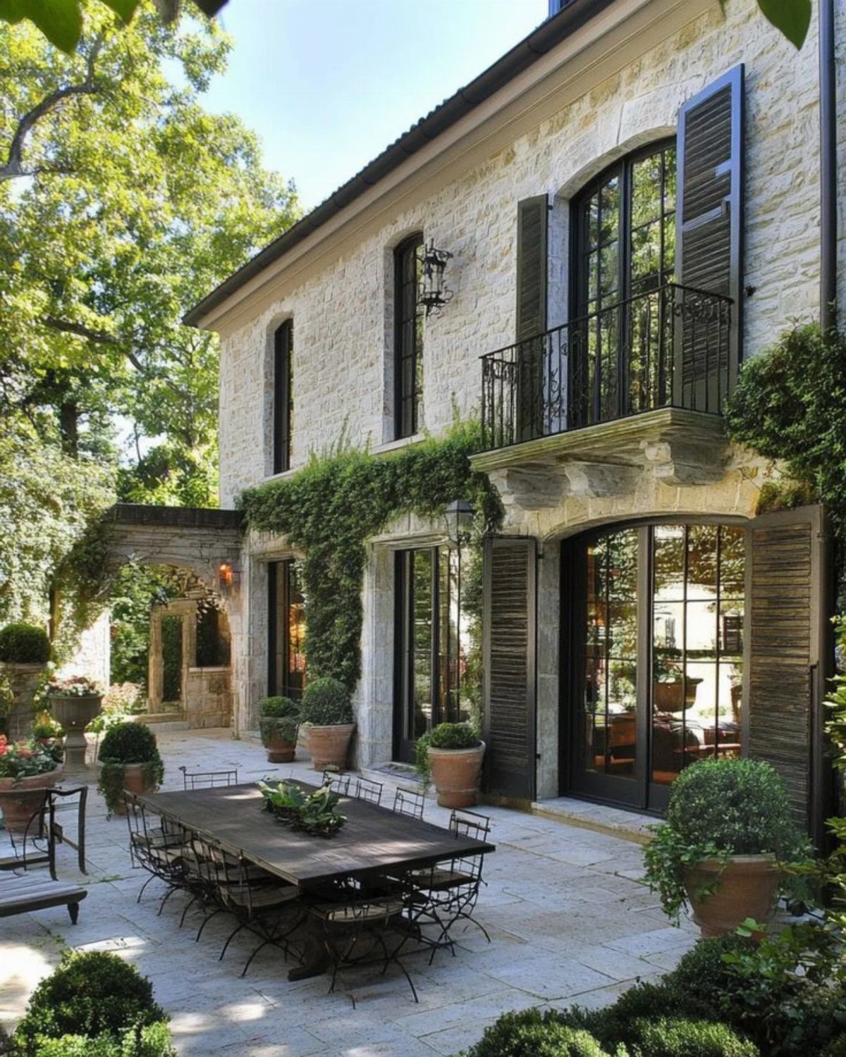 A stone patio with a large wooden dining table and black chairs surrounded by potted greenery.
