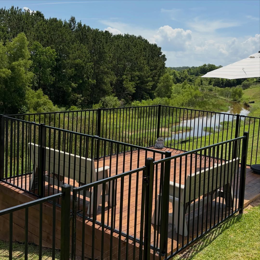 A reddish-brown wooden deck with black metal railings overlooking a green forest and stream.