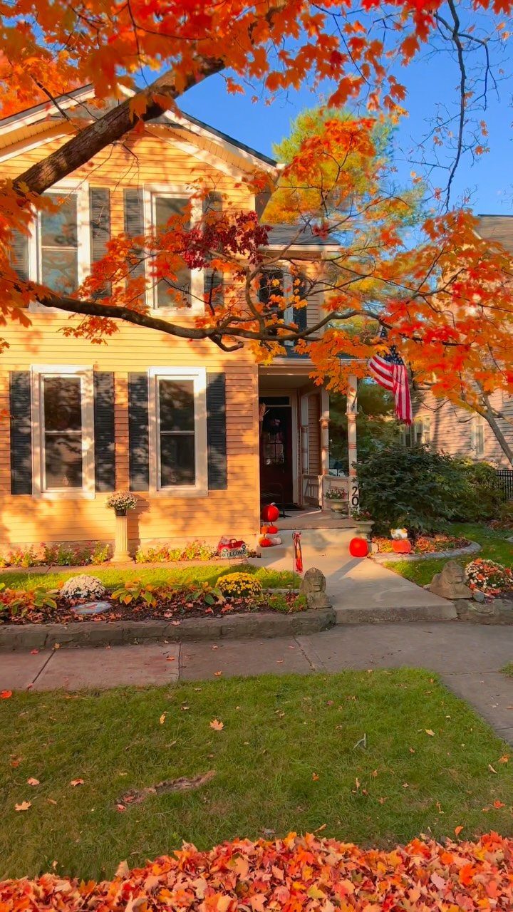 12 Trending House Colors for Exterior Looks in 2026 12 A tan two-story house with dark shutters framed by bright orange autumn leaves.