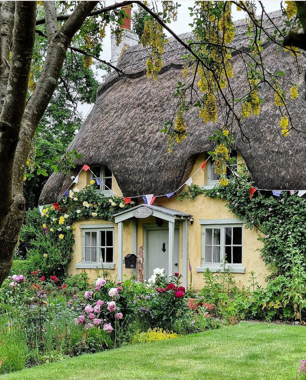 A yellow thatched cottage with green window frames surrounded by a blooming flower garden.