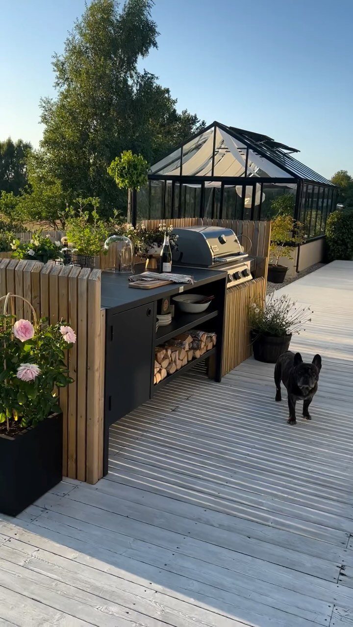 10 Wooden Garden Kitchen Ideas for Warm Outdoor Cooking 6 Outdoor kitchen island with black countertop, firewood storage, and a stainless steel grill on a wooden deck.